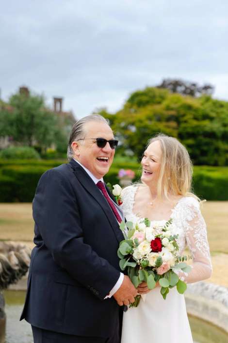 joyful outdoor wedding moment A smiling bride in a lace wedding dress holds a bouquet of roses and greenery while standing with a man in a dark suit and sunglasses outdoors.