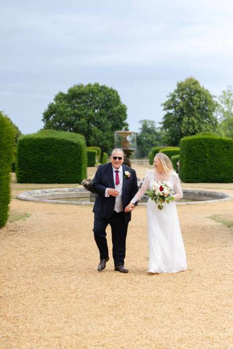 bride and father walking in garden Bride in a white wedding gown carries a bouquet as she walks hand in hand with a man in a dark suit along a gravel garden path, with trimmed hedges and a fountain in the background.