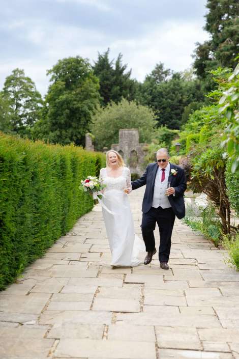 bride and father walking garden path Bride in a white wedding dress walks along a stone garden path, holding a bouquet, with a man in a dark suit beside her. Photo by Louisa Dettmer Wedding Photography, Kent Wedding Photographer