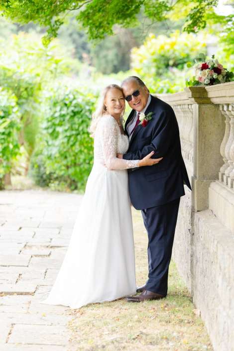 romantic garden wedding portrait A bride in a white lace wedding dress embraces a man in a dark suit beside a stone railing with a bouquet in a lush garden. Photo by Louisa Dettmer Wedding Photography, Kent Wedding Photographer