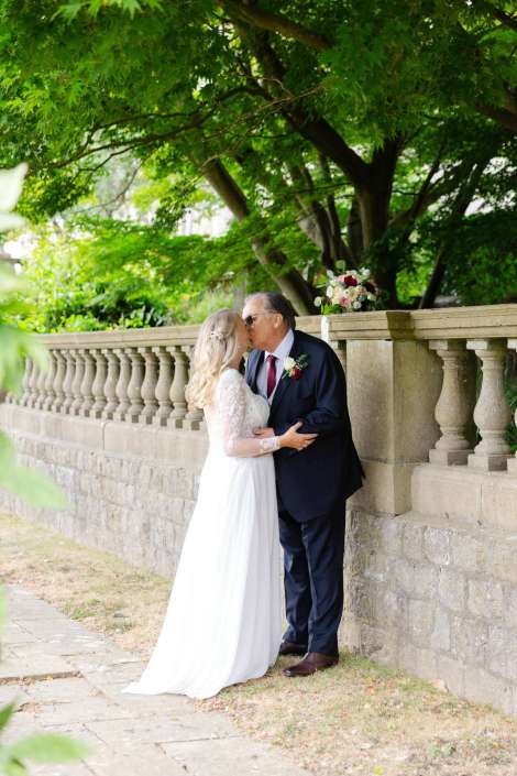 bride and groom kiss outdoors Bride in a white lace wedding dress shares a kiss with an older man in a dark suit beside a stone balustrade under leafy trees, with a bouquet resting on the wall. Photo by Louisa Dettmer Wedding Photography, Kent Wedding Photographer.