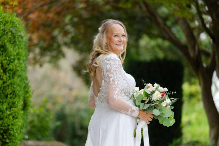 bride in lace wedding dress Bride in a white lace wedding dress holds a bouquet and smiles outdoors in a lush garden. Photo by Louisa Dettmer Wedding Photography, Kent Wedding Photographer