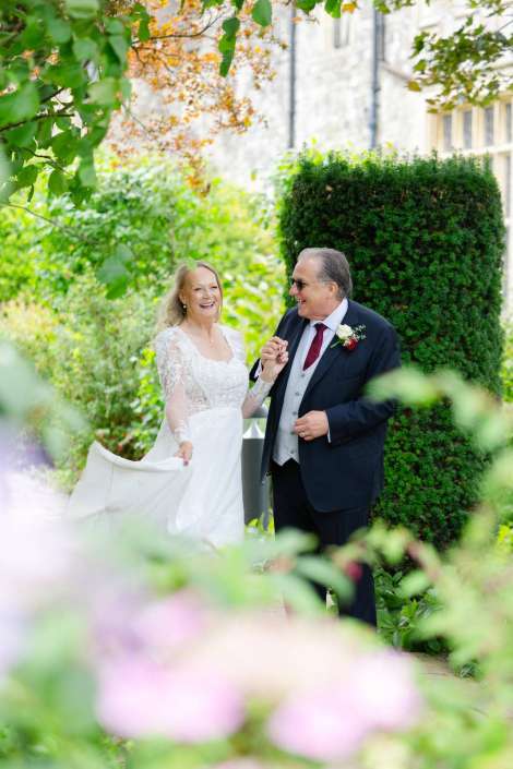 bride and father dancing in garden A bride in a white lace wedding dress walks hand in hand with a man in a dark suit through a lush garden, both smiling. Photo by Louisa Dettmer Wedding Photography, Kent Wedding Photographer