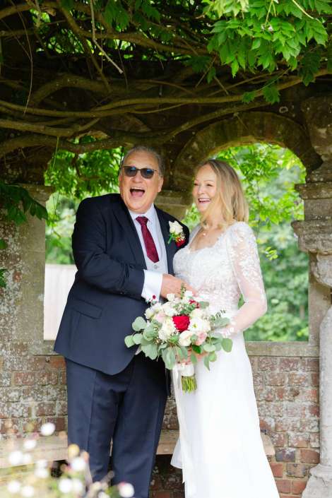 joyful wedding couple portrait A bride in a white lace gown and a groom in a dark suit laugh together under a leafy stone arch, the bride holding a bouquet.