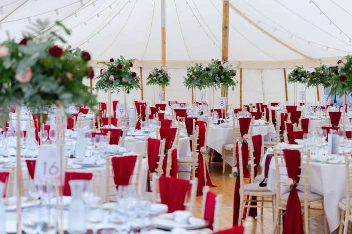 elegant tent wedding reception A wedding reception inside a large white tent with round tables dressed in white linens, red napkins, and tall floral centerpieces.