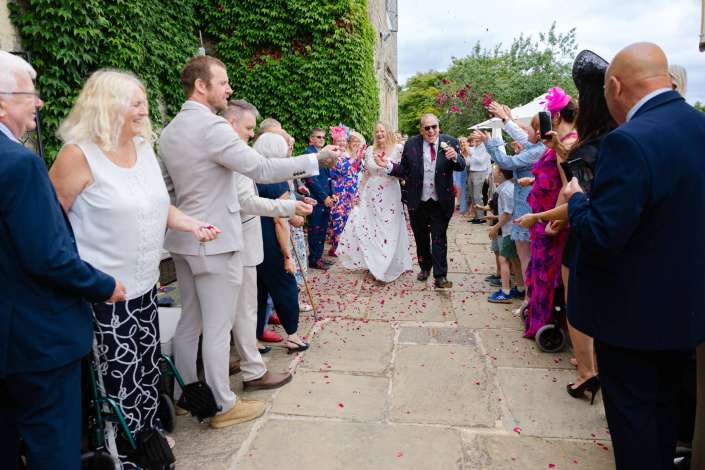 bride and groom confetti sendoff Bride in a white gown and groom in a dark suit walk down a petal-strewn stone path as wedding guests on both sides shower them with pink petals.