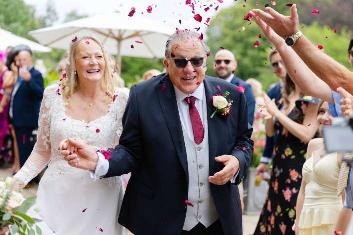 confetti shower at wedding A bride in a white lace wedding dress walks arm-in-arm with an older man in a dark suit as pink confetti falls at an outdoor wedding.