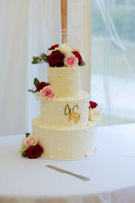 elegant three tier wedding cake Three-tier white wedding cake adorned with small pearls, a gold G&S monogram, and red, pink, and white roses, placed on a white table with a cake knife nearby.