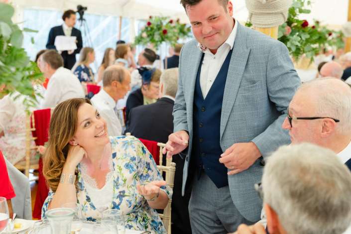 joyful conversation at wedding reception A man in a gray plaid suit chats with a smiling woman in a floral dress at a crowded banquet table during a celebration.