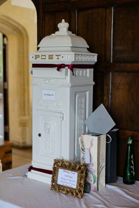 wedding card box display A tall, ornate white mailbox-style wedding card box with a domed lid and burgundy ribbon sits on a table beside gift bags.