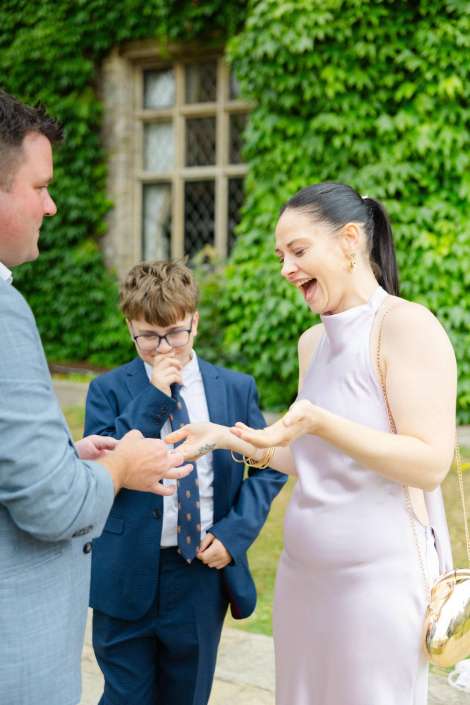 joyful outdoor wedding moment A woman in a pale pink dress laughs as a man in a suit places a ring on her finger, while a boy in a blue suit watches nearby.