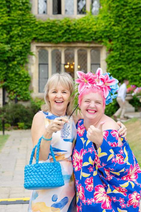 joyful friends at garden party Two women pose outdoors in front of an ivy-covered building; one wears a blue tropical dress with a large pink flower headpiece, while the other wears a pastel dress and carries a bright blue woven bag while holding a drink.