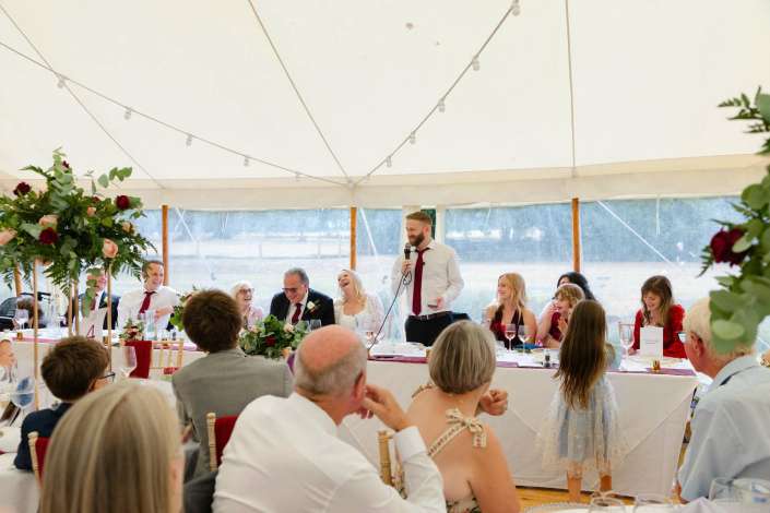 wedding reception speech under tent Wedding reception under a white tent with string lights, guests seated at long tables with floral arrangements while a man gives a speech.