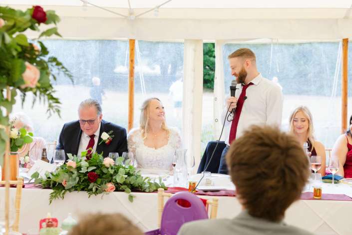 wedding speech under tent Bride and groom sit at a flower-decorated wedding reception table inside a tent as a man in a white shirt and red tie speaks into a microphone.
