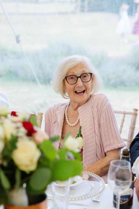 joyful elderly woman at table Smiling elderly woman with white hair and glasses, wearing a pink cardigan and pearl necklace, seated at a table with a floral centerpiece.