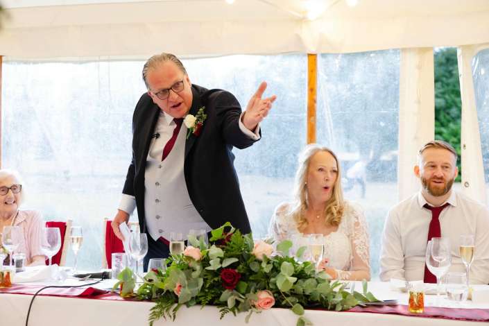toast at wedding reception A man in a suit delivers a toast at a wedding reception, gesturing toward guests seated at a flower-decorated head table with the bride nearby.
