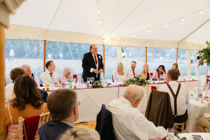 wedding speech under string lights A man in a suit giving a toast at a wedding reception inside a white tent, with guests seated around a long table under string lights.