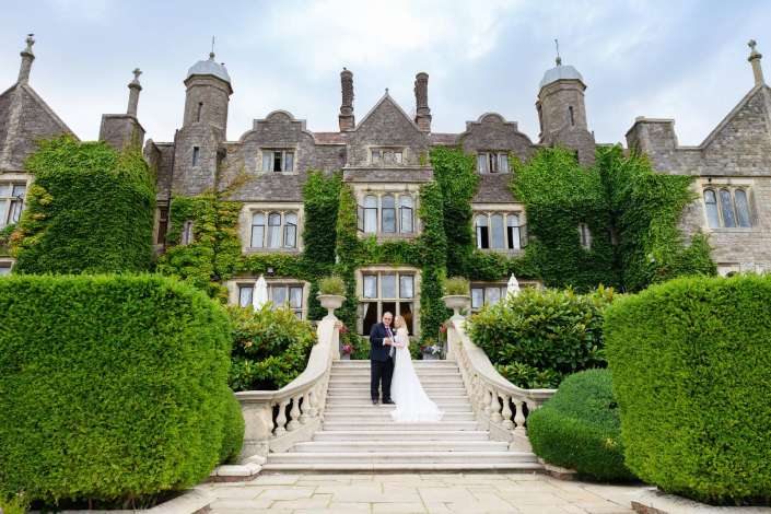 ivied castle wedding Bride and groom stand on a grand stone staircase in front of an ivy-covered historic manor with towers. Photo by Louisa Dettmer Wedding Photography, Kent Wedding Photographer