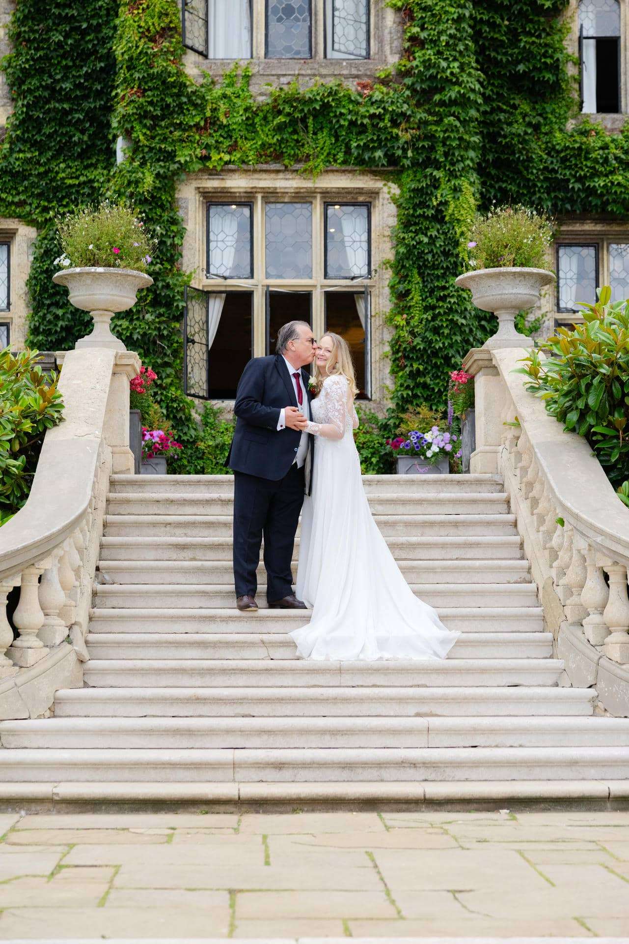ivy covered staircase wedding kiss Bride in a white gown and groom in a dark suit kiss on a grand stone staircase outside an ivy-covered building with flowering planters. Photo by Louisa Dettmer Wedding Photography, Kent Wedding Photographer