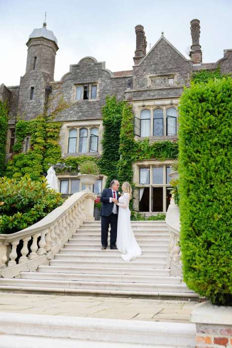 castle staircase wedding portrait A bride in a white wedding gown and a groom in a dark suit stand on a grand stone staircase in front of an ivy-covered historic mansion. Photo by Louisa Dettmer Wedding Photography, Kent Wedding Photographer.