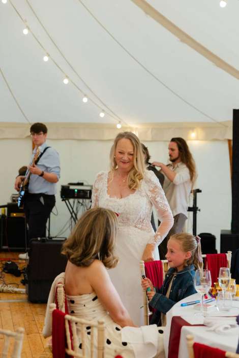 bride socializing with guests Smiling bride in a lace wedding gown chats with two guests at a table as a live band plays under string lights inside a white tent.