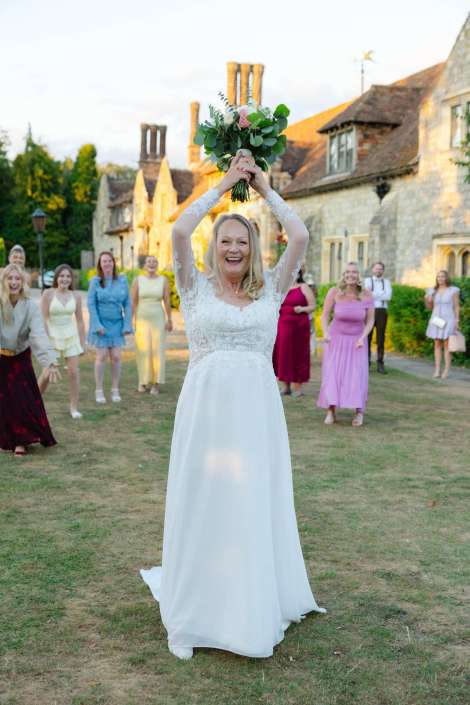 bridal bouquet raised high Bride in a white lace wedding gown raises a bouquet above her head and smiles, while bridesmaids in pastel dresses stand on the lawn behind her.
