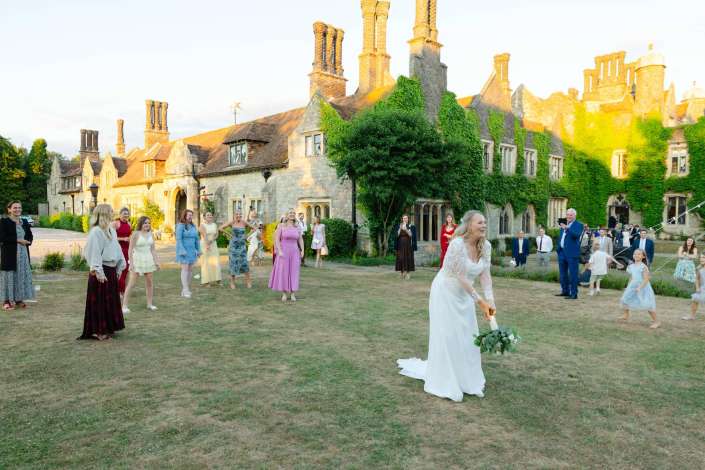 sunlit ivy manor wedding Bride in a white wedding dress with a bouquet stands on a lawn as guests in colorful dresses mingle outside an ivy-covered stone manor.