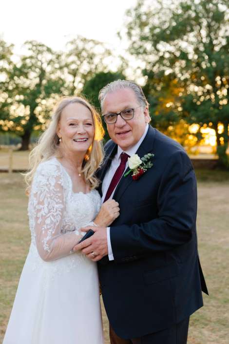 joyful outdoor wedding portrait A smiling bride in a lace long-sleeve wedding dress and a groom in a dark suit with a boutonniere stand together outdoors at sunset.