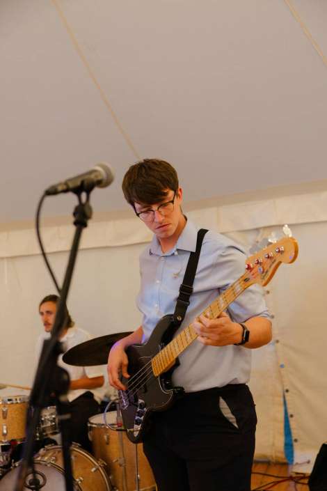 bassist on stage A young man in a light blue shirt and glasses plays a black bass guitar on stage, with a drummer in the background. Photo by Louisa Dettmer Wedding Photography, Kent Wedding Photographer