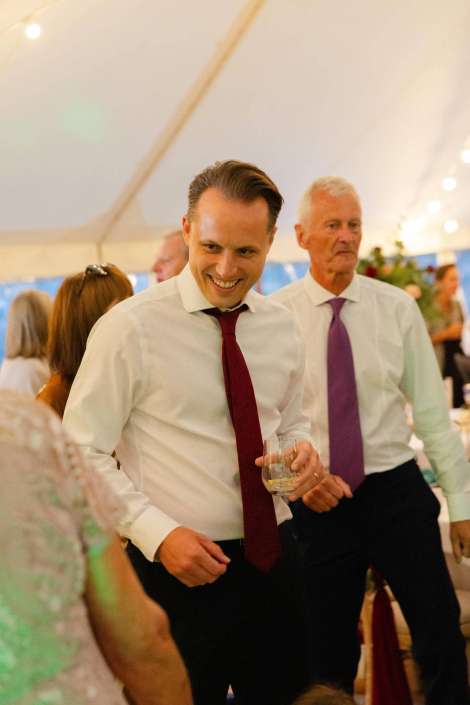smiling man at reception A smiling man in a white shirt and red tie holds a glass at a wedding reception inside a tent. Photo by Louisa Dettmer Wedding Photography, Kent Wedding Photographer