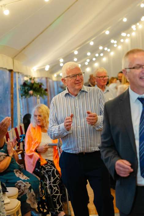 man in striped shirt clapping An older man with glasses in a blue-striped shirt claps as he walks past seated guests at a wedding reception inside a string-lit tent. Photo by Louisa Dettmer Wedding Photography, Kent Wedding Photographer.