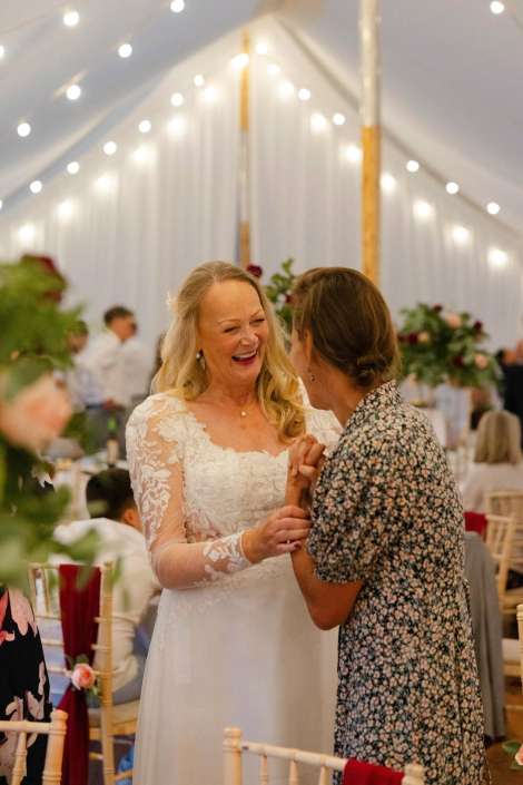 joyful bride with relative Bride in a white lace wedding dress laughs while holding hands with a guest in a floral dress at a softly lit wedding reception. Photo copyright Louisa Dettmer Wedding Photography