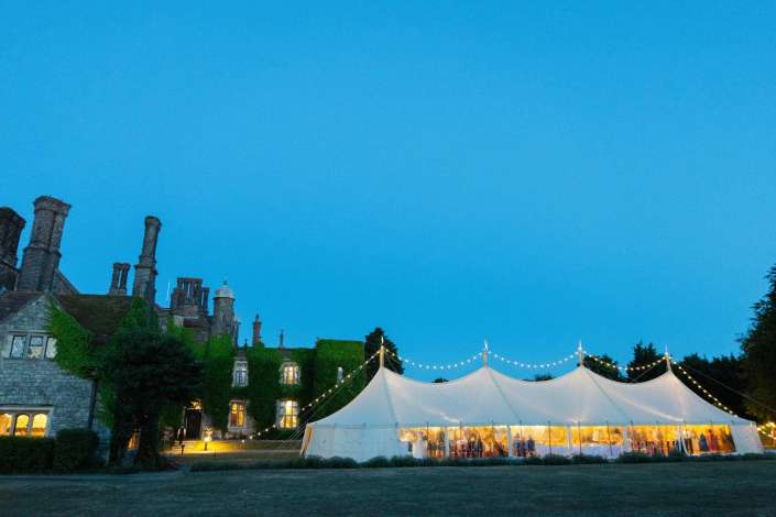 castle courtyard tent at night A historic stone building with tall chimneys and ivy sits beside a lit white wedding marquee under a deep blue evening sky. Photo by Louisa Dettmer Wedding Photography, Kent Wedding Photographer