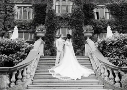 A bride in a long wedding gown and a groom in a light suit stand on a grand stone staircase outside an ivy-covered building, with the bride's train trailing down the steps.