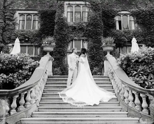 A bride in a long wedding gown and a groom in a light suit stand on a grand stone staircase outside an ivy-covered building, with the bride's train trailing down the steps.