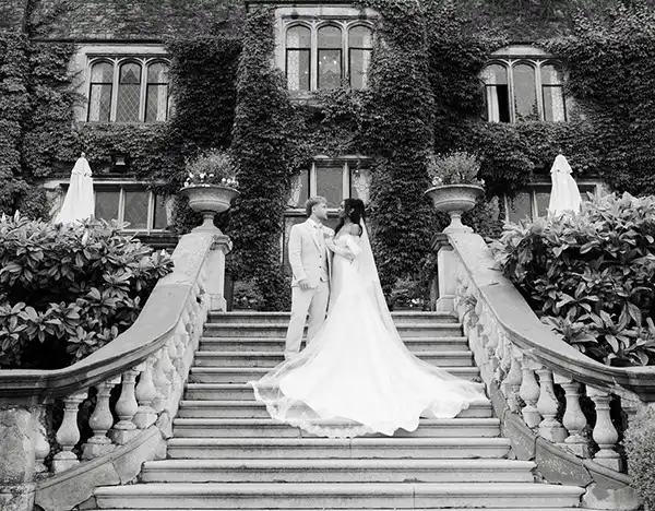 A bride in a long wedding gown and a groom in a light suit stand on a grand stone staircase outside an ivy-covered building, with the bride's train trailing down the steps.