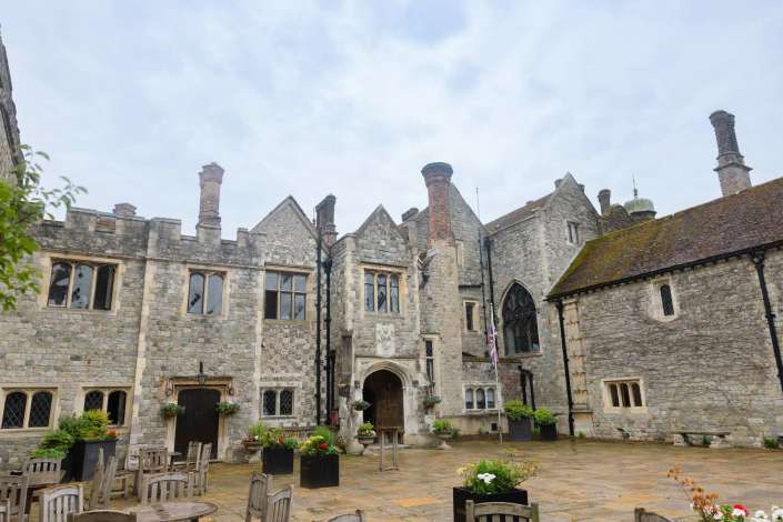 historic stone courtyard A historic stone building with arched windows and tall chimneys surrounds a paved courtyard lined with wooden tables, chairs, and potted flowers.