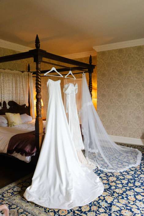 bridal gowns on four-poster bed Three white wedding dresses on hangers beneath a dark four-poster bed in a luxurious bedroom with a blue floral carpet and a long lace veil trailing on the floor.
