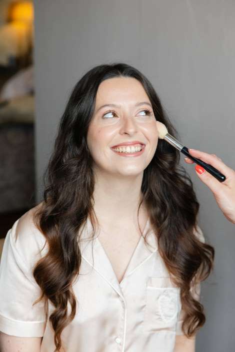 smiling woman getting makeup Smiling woman with long, wavy dark hair wearing a pale satin pajama top while a makeup brush applies makeup to her cheek.