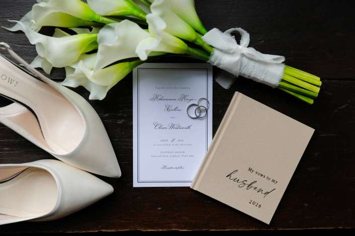 bridal bouquet with rings Flat lay of wedding essentials on a dark wooden surface: ivory heels, a white calla lily bouquet tied with ribbon, a wedding invitation with rings, and a beige guest book.