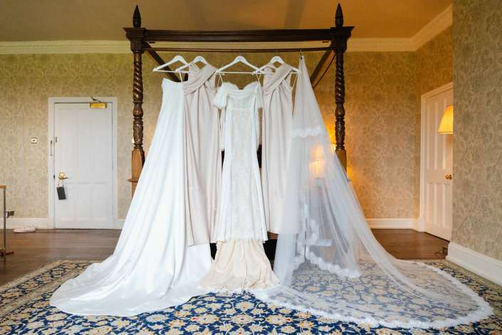 bridal gowns on canopy bed Bridal gowns hang from a dark wooden four-poster bed in a vintage room with floral wallpaper and a blue patterned rug.