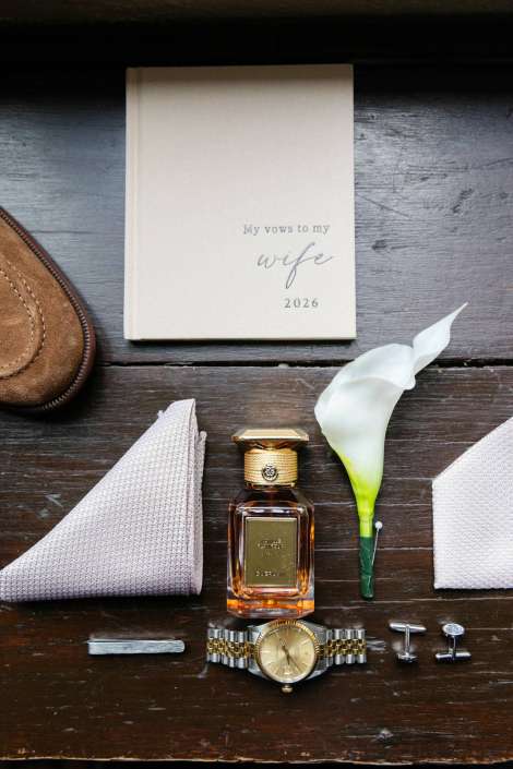 vows to my wife 2026 Flat lay of wedding accessories on a dark wooden surface, including a beige vow book, a perfume bottle, a white calla lily, a gold-and-silver wristwatch, folded napkins, cufflinks, and a brown leather shoe.