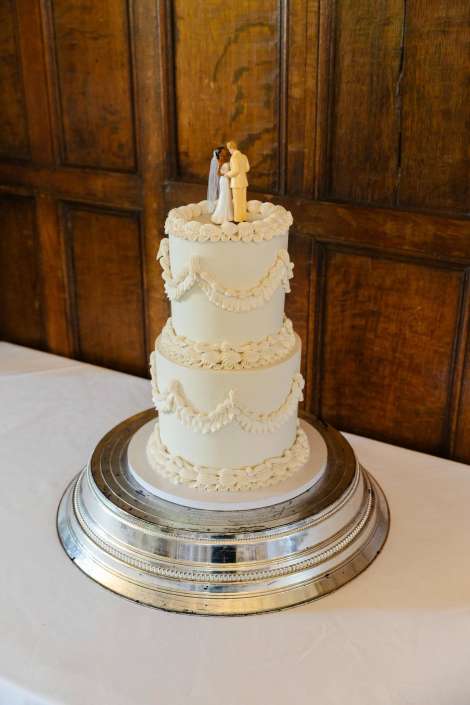 ivory three tier wedding cake Three-tier white wedding cake with decorative piped borders and swags, topped by a bride and groom figurine, placed on a silver stand.