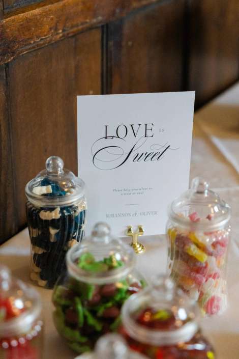 love is sweet card display A white card with elegant script reading “LOVE is Sweet” stands on a table beside glass jars filled with colorful candies.