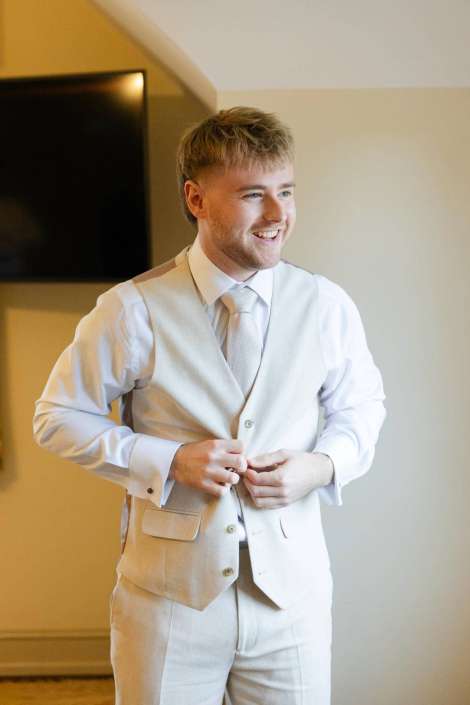 groom buttoning cream suit Smiling man in a light beige suit and tie buttons his vest indoors.