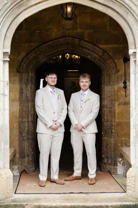 two grooms in beige suits Two men in matching light beige suits with boutonnieres stand side by side in a stone church doorway.