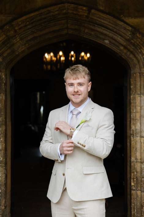 groom in light beige suit A man in a light beige suit stands in a stone arched doorway, adjusting his cuff while wearing a white boutonniere.