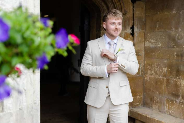 groom in beige suit Smiling man in a light beige suit and pale pink tie stands in a stone archway with purple flowers in the foreground.