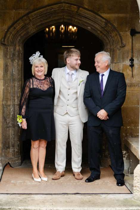groom with his parents outside church Groom in a light beige suit stands with a woman in a black lace dress and a man in a dark suit under a stone arched doorway, all smiling.