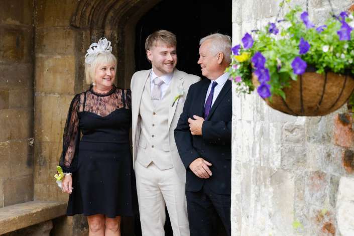 wedding party at stone arch Three adults in formal attire smile together in a stone archway, with a hanging basket of purple flowers on the right.